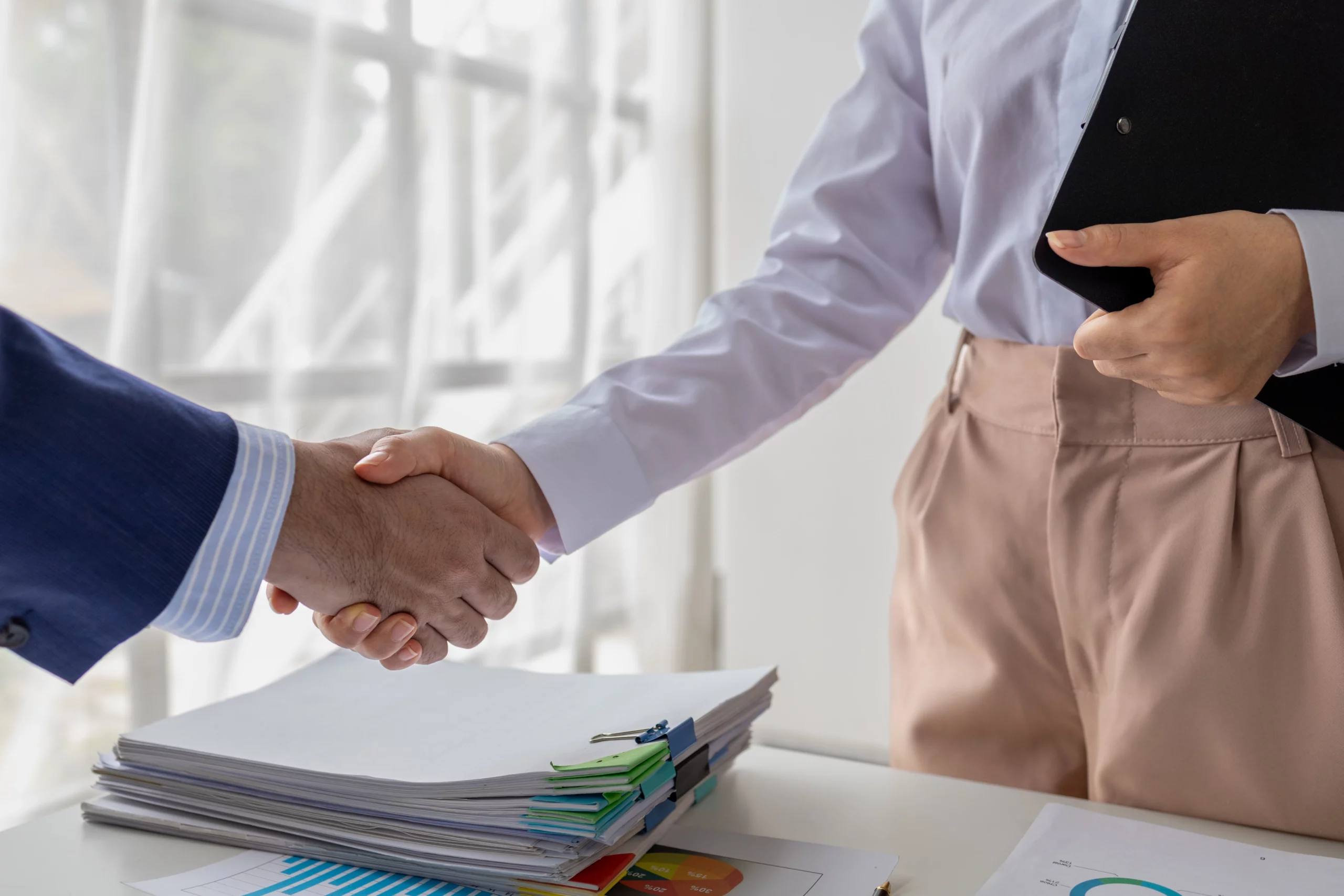 Two people shake hands over documents and a clipboard, symbolizing a mutual agreement reached during divorce mediation in a professional setting.