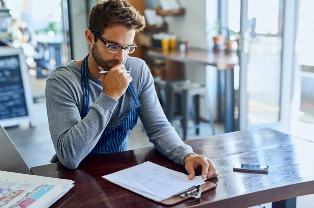 Small business owner in apron sits at a café table, thoughtfully reviewing papers on a clipboard, with a smartphone and newspaper beside him.
