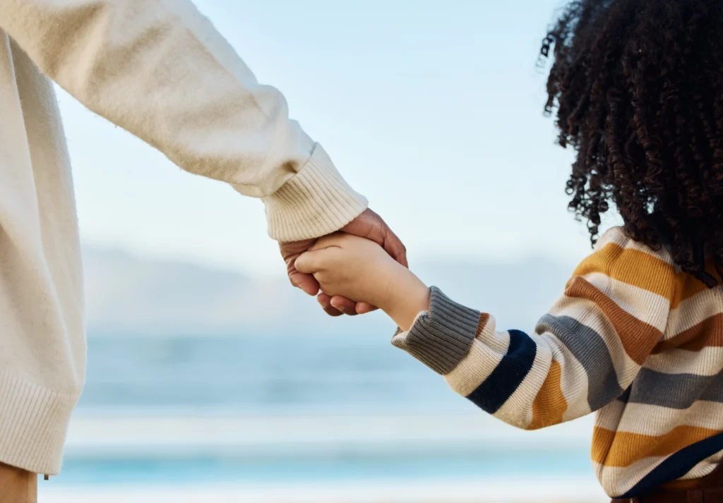 Close-up of an adult and child holding hands by the beach, child in a striped sweater; symbolizes care, stability, and parent-child connection in custody matters.