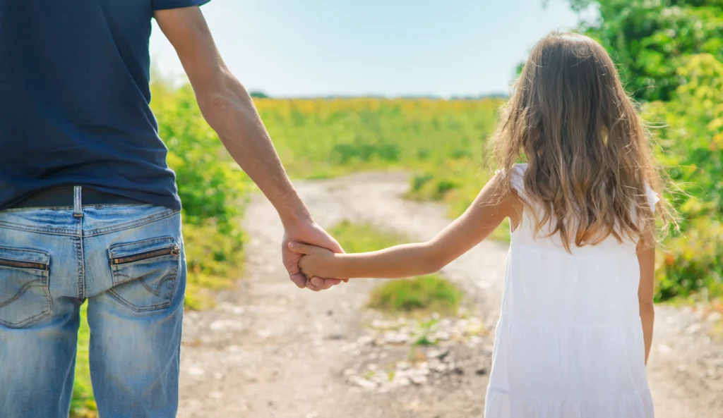 Man and young girl walking hand in hand along a sunny path through green fields, representing the importance of parent-child relationships and the best interests of the child in child custody decisions in Sonoma County, California.