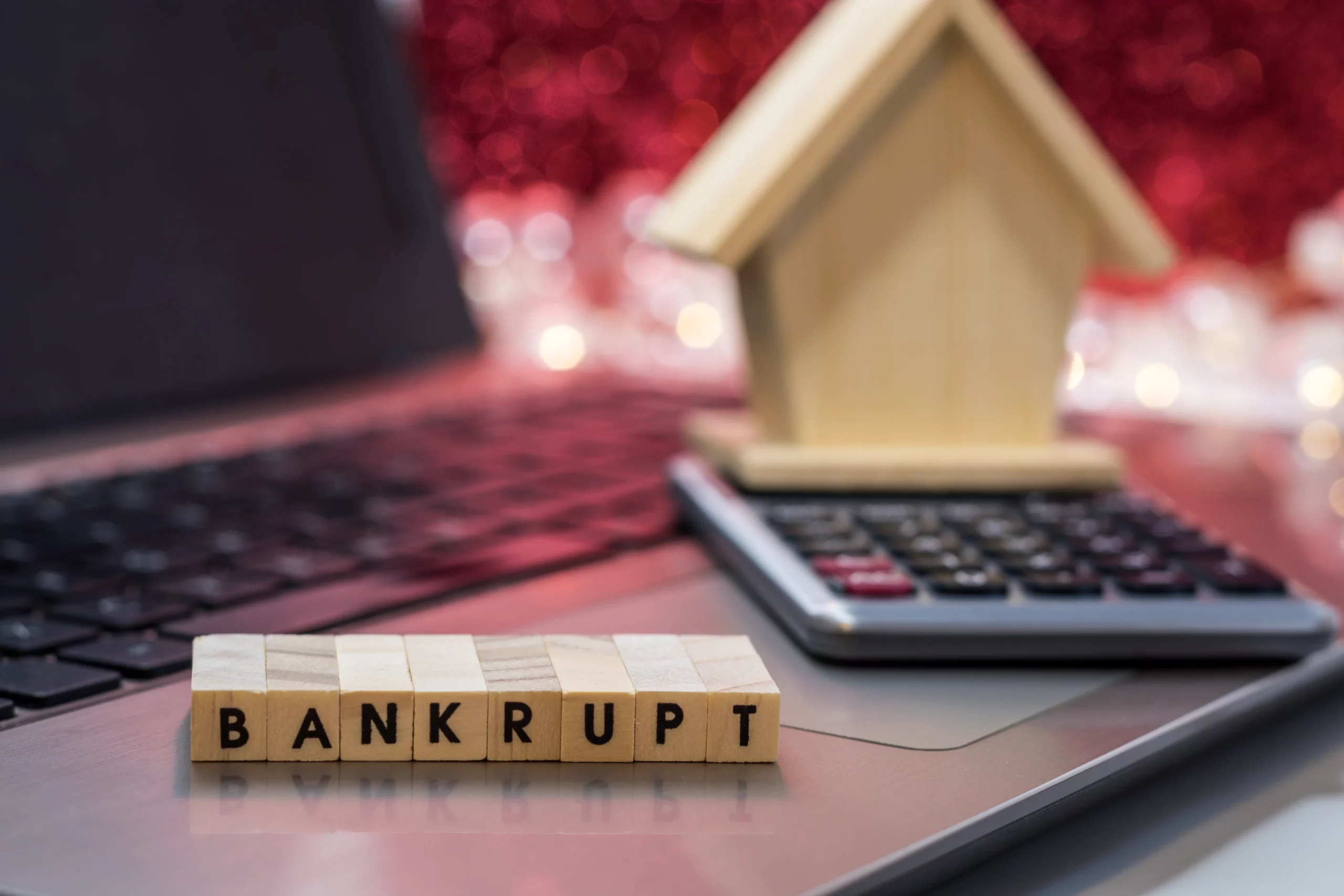 Wooden blocks spelling “BANKRUPT” placed on a laptop beside a model house and calculator, representing financial hardship and explaining which debts may still remain after Chapter 7 bankruptcy in California.